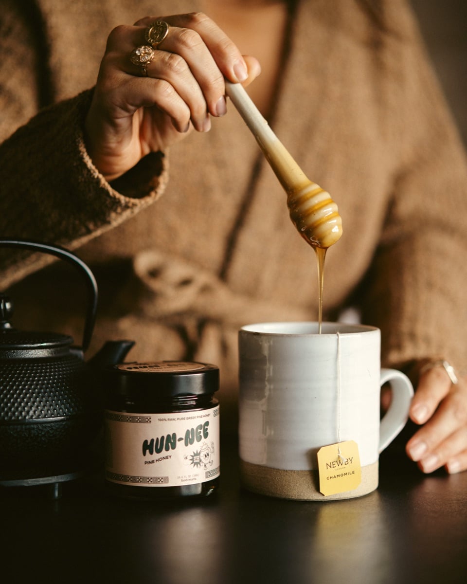 Honey being poured into a mug beside Hun-Nee jars.