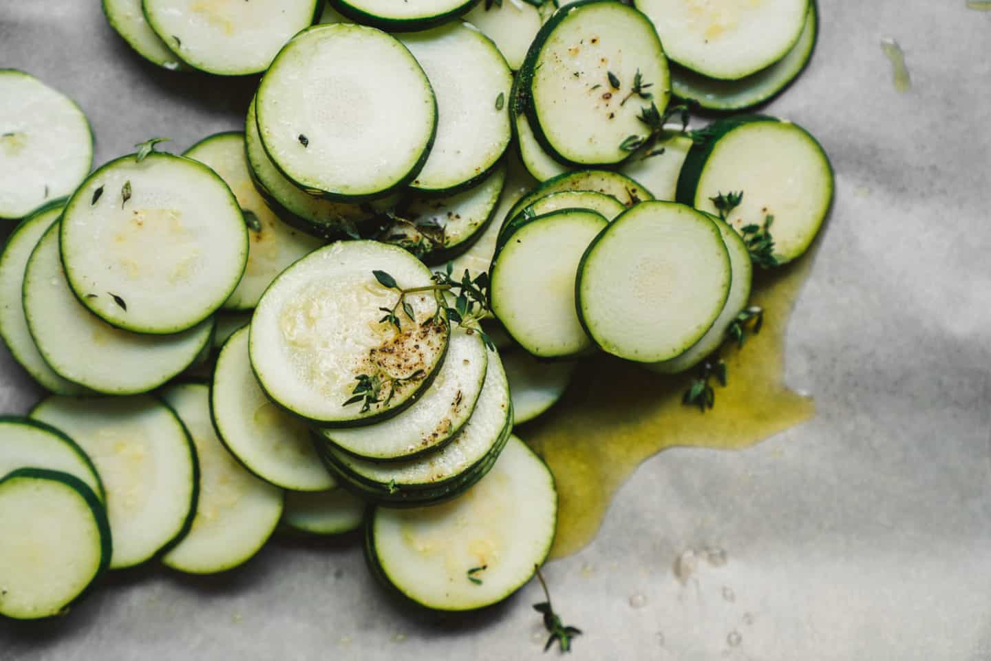 Thinly sliced zucchini rounds arranged on a parchment-lined baking sheet, drizzled with olive oil and sprinkled with fresh thyme and seasonings