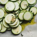 Thinly sliced zucchini rounds arranged on a parchment-lined baking sheet, drizzled with olive oil and sprinkled with fresh thyme and seasonings