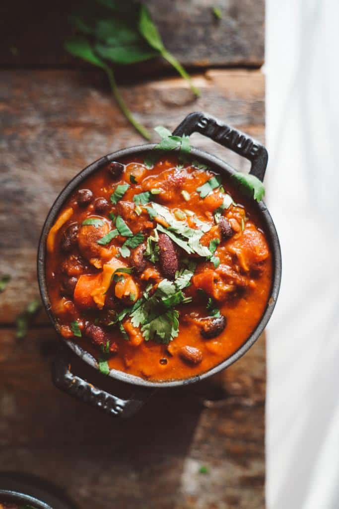 A pot of rich, red chili on a wooden surface, garnished with fresh cilantro. The chili contains beans and vegetables, adding texture and color. Bright natural light highlights the dish, with a sprig of cilantro nearby.