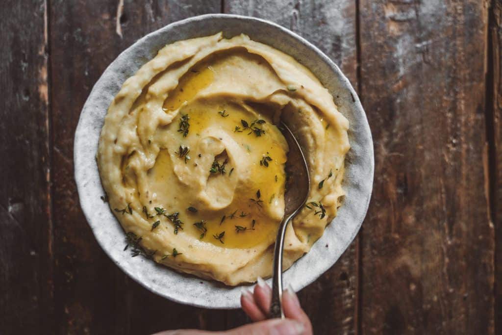 A bowl of creamy onion garlic mashed potatoes topped with melted butter and garnished with herbs. A hand holds a spoon resting in the bowl, placed on a rustic wooden table.