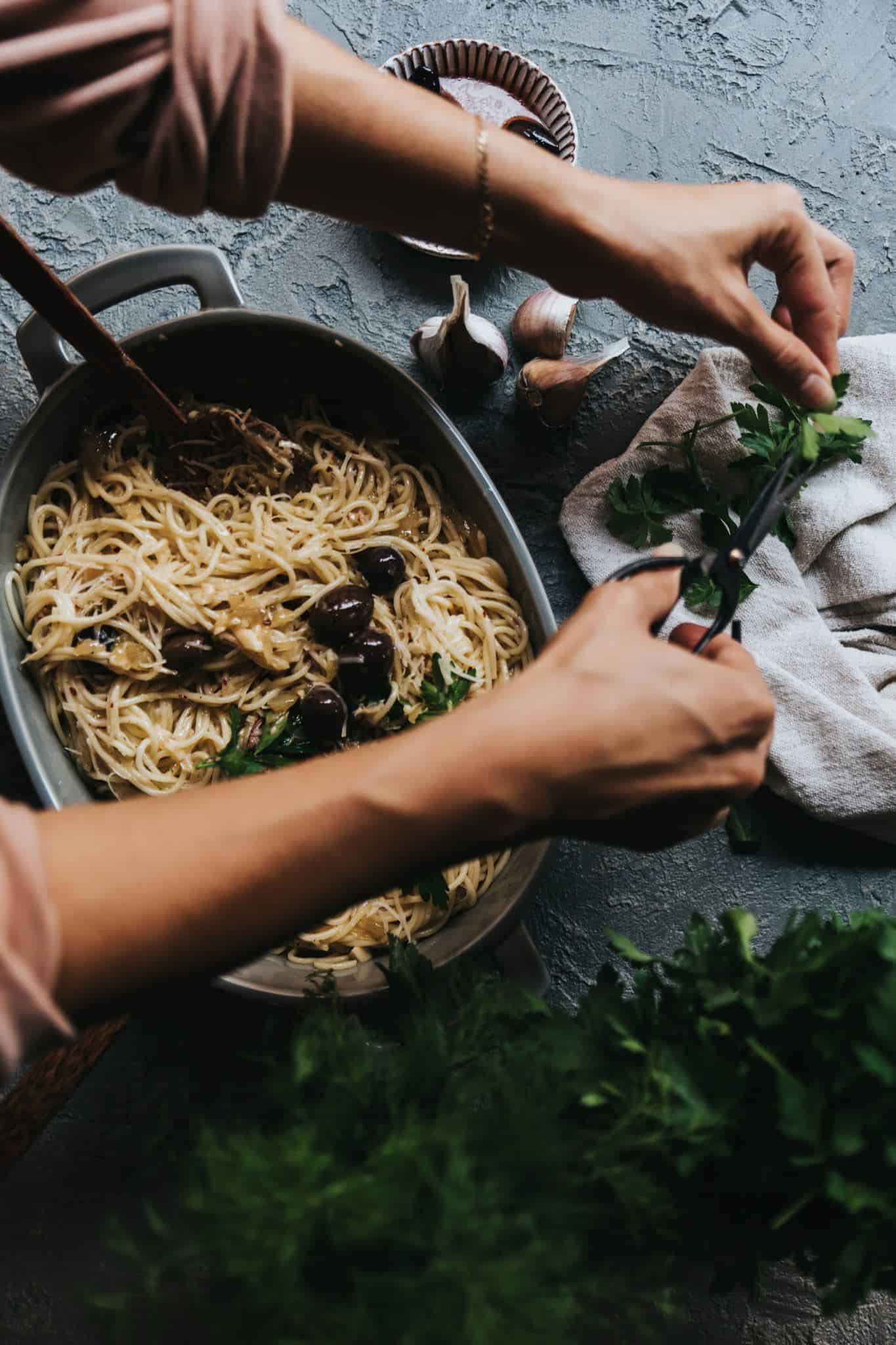 Creamy Vegan Pasta in a large serving dish with arms reaching over to cut fresh herbs for garnish.