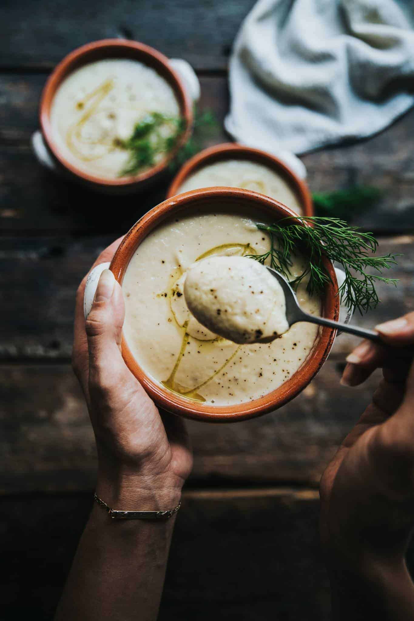 A hand holding a bowl of cauliflower soup with a big piece of fresh dill on top and  a spoon scooping some out