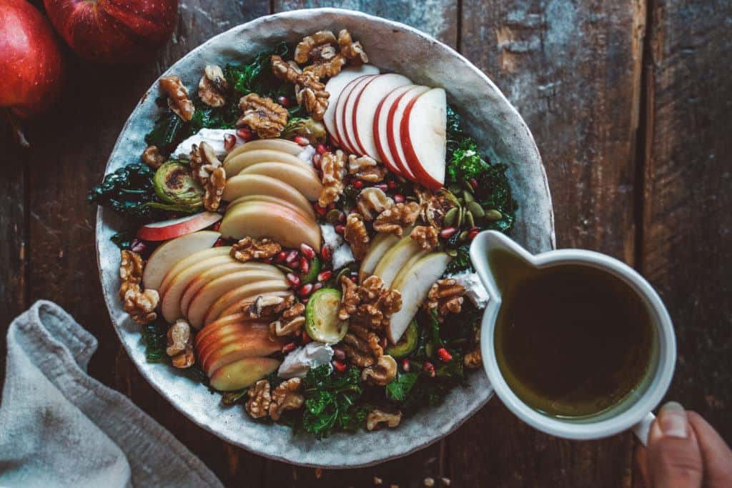 A rustic bowl filled with sliced apples, walnuts, pomegranate seeds, and kale. A person is pouring a dressing from a small pitcher onto the salad. A red apple and a napkin are nearby on a wooden table.