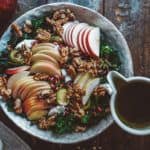 A rustic bowl filled with sliced apples, walnuts, pomegranate seeds, and kale. A person is pouring a dressing from a small pitcher onto the salad. A red apple and a napkin are nearby on a wooden table.