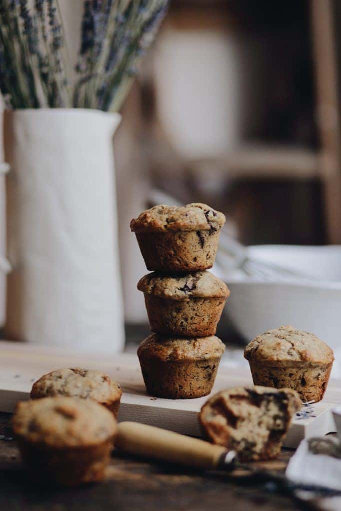 A stack of three muffins on a wooden board with two more muffins on the surface. A white vase with lavender stems and two white bowls are blurred in the background. A knife with crumbs is on the board.