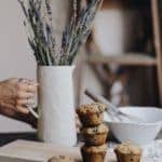 A person holding a white vase with lavender stems next to a stack of muffins on a wooden cutting board. A white bowl and a whisk are in the background, with a ladder leaning against the wall.