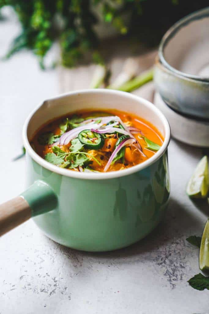 A green pot filled with vibrant soup topped with sliced red onions, jalapeños, and fresh cilantro. The soup is a rich orange color. In the background are stacked bowls and lime wedges. The setting is a light, rustic kitchen surface.