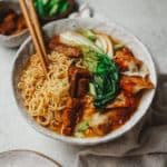 A bowl of ramen noodles with bok choy, dumplings, and sliced meat, accompanied by chopsticks. A small bowl of greens and another with dumplings are in the background on a neutral cloth-covered surface.