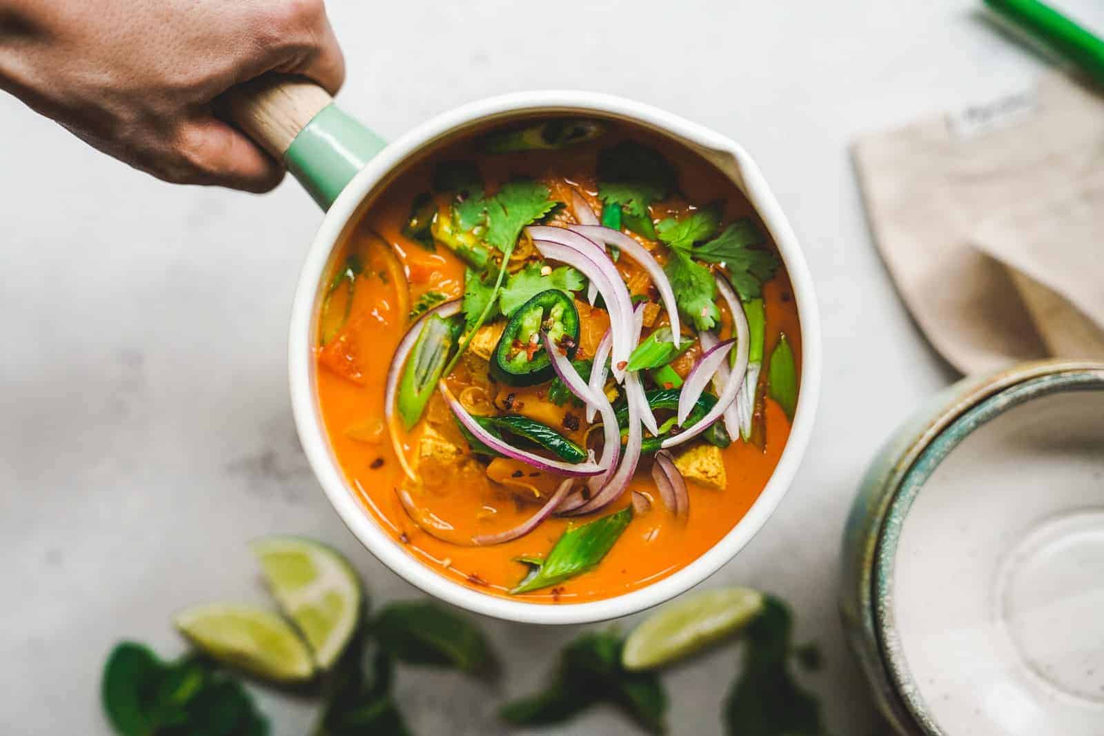 A pot with bright orange curry with toppings of fresh herbs and red onion. 