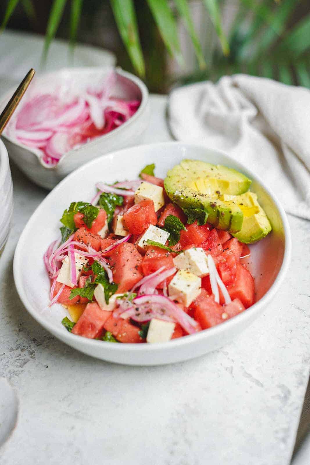 Bowl of fresh watermelon, vegan feta, and avocado on white table with pickled red onions in the background.