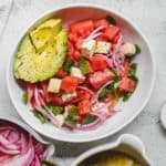 A bowl of refreshing watermelon mint feta salad features watermelon cubes, sliced avocado, feta cheese, red onion, and fresh mint. Alongside the bowl are lime halves, ground pepper, and a dish of sliced red onions on a light surface.