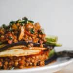 A close-up of a Vegan Moussaka dish featuring layered roasted vegetables and lentils in a rich tomato-based sauce, garnished with chopped herbs, served on a white plate against a neutral background.