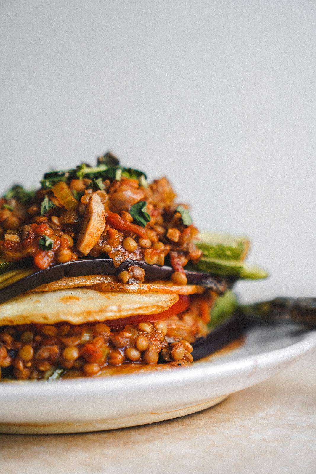 A close-up of a Vegan Moussaka dish featuring layered roasted vegetables and lentils in a rich tomato-based sauce, garnished with chopped herbs, served on a white plate against a neutral background.
