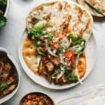 A plate features a vegan gyro with grilled plant-based meat, lettuce, tomatoes, onions, and herbs on flatbread. Surrounding bowls offer roasted vegetables and diced tomatoes for added flavor.