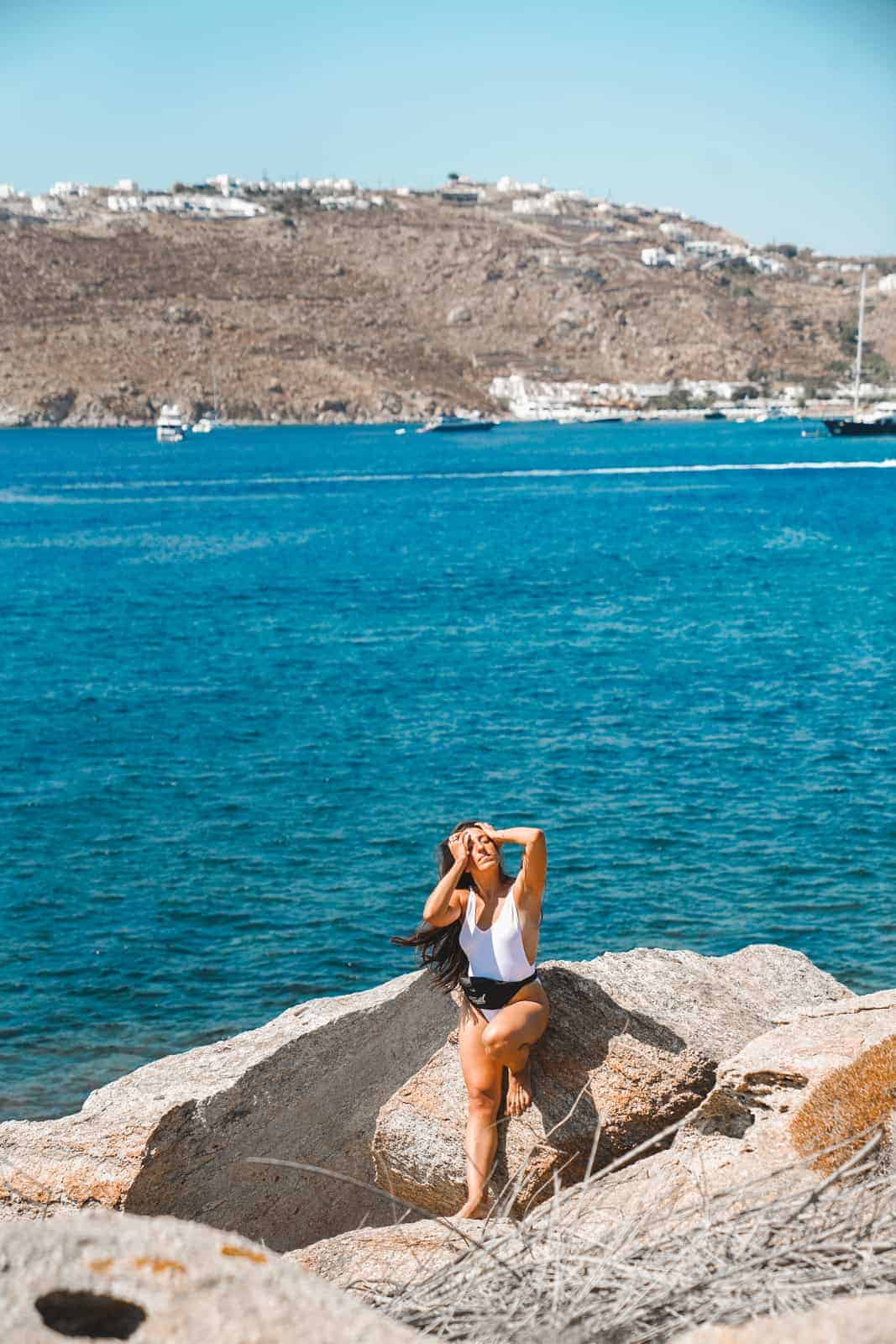 Maria sitting on a rock by the ocean in Mykonos, Greece. Ready to give you her international travel tips!
