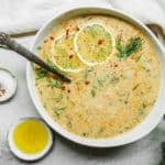 A bowl of vegan Avgolemono soup, creamy and garnished with lemon slices, fresh dill, and a sprinkle of red pepper flakes. A spoon rests inside the bowl. Nearby are small bowls of salt and olive oil, a glass of water with lemon, and a white cloth.
