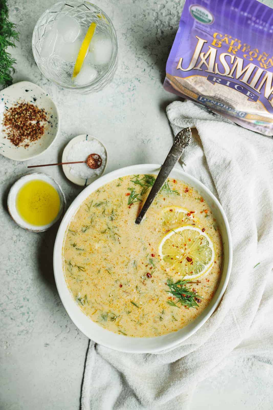 A big bowl of Greek Lemon Rice Soup on counter surrounded by key ingredients. An amazing vegan meal!