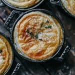 Close-up of mini pot pies in black dishes on a wooden surface, their golden, flaky crusts topped with fresh thyme. These Cream of Mushroom Pot Pies evoke a rustic, home-cooked charm that warms the soul.