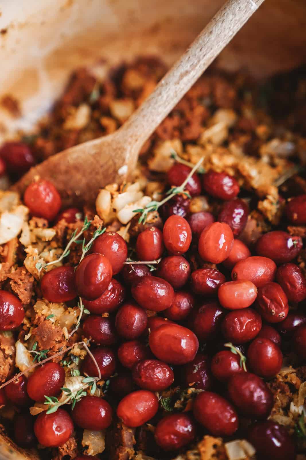 A wooden spoon in a dish of cooked stuffing mixed with fresh red cranberries, onions, and herbs, creating a colorful and textured appearance.