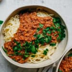 A bowl of vegan spaghetti topped with rich, hearty meat sauce and garnished with fresh parsley. The dish is placed on a light-colored surface, accompanied by additional parsley leaves visible nearby.