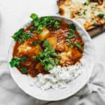 A bowl of curry with potatoes and herbs, served alongside white rice. The dish is garnished with fresh cilantro. In the background, there is a piece of naan bread topped with herbs, all placed on a light-colored surface.