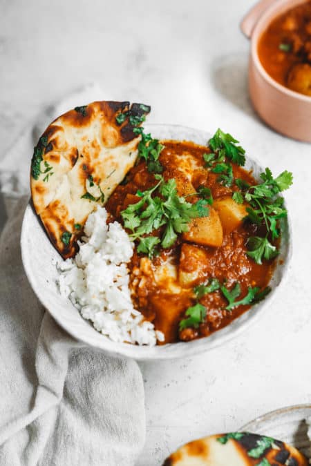 A bowl of curry with chunks of potato, garnished with cilantro, accompanied by white rice and a piece of naan bread. The dish is served in a white bowl on a light-colored surface, with a soft fabric napkin next to it.