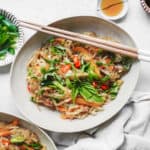 A bowl of Vegan Thai Drunken Noodles is topped with fresh basil, red chili slices, and vibrant vegetables. Chopsticks rest on the bowls edge. Nearby are a small dish of green onions and two small bowls with sauces. A light cloth is partially visible.