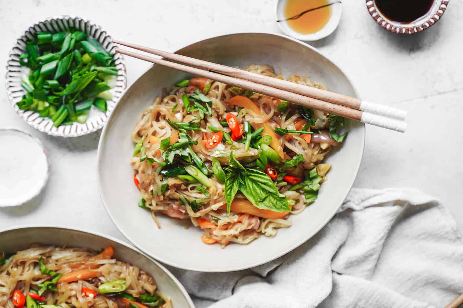 White bowl filled with this drunken noodles recipe with chopsticks surrounded by fresh ingredients