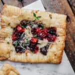 A square puff pastry topped with cranberry cheese and roasted cherry tomatoes rests on a white marble surface. The golden brown vegan tart is flaky, sprinkled with green herbs, and set against a rustic wooden background.