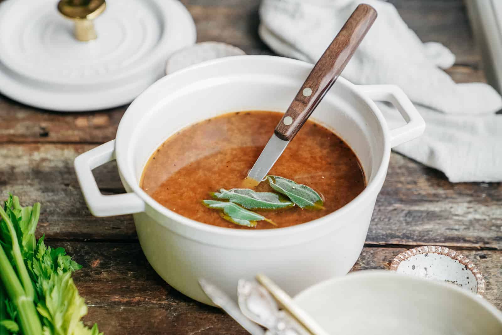 Vegetable broth in a pot with a spoon and fresh herbs on top.