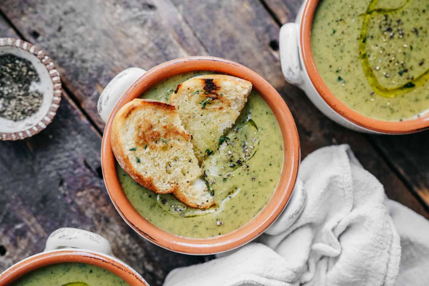 A bowl of Cream of Broccoli Soup, garnished with toasted bread slices sits on a wooden surface. Nearby, a small bowl contains ground pepper, and a folded white cloth lies beside the soup. Two additional bowls of this delightful soup are partially visible.