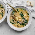 A bowl of creamy vegan gnocchi soup with mushrooms and spinach, garnished with herbs. A spoon rests in the bowl. Pieces of cheese alternatives are scattered nearby. A cloth napkin is placed beside the bowl on a light-colored surface.
