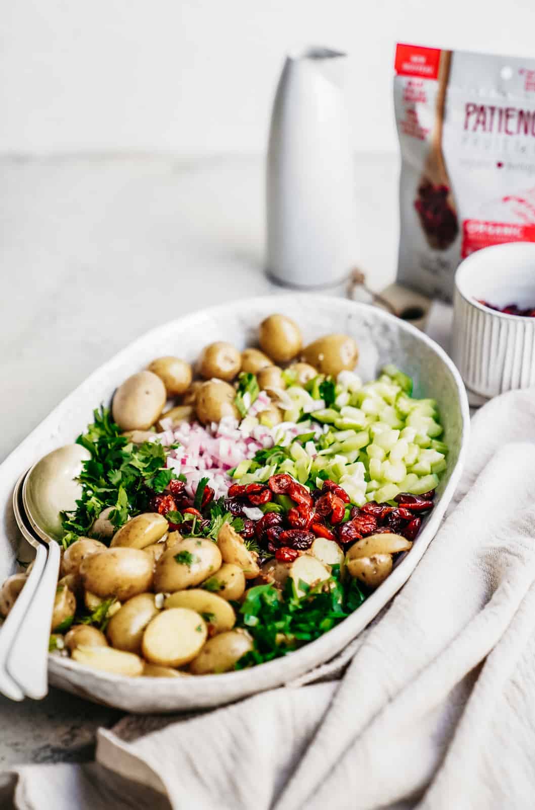 A bowl of Vegan Potato Salad in a serving dish with a bag of dried cranberries in the back.