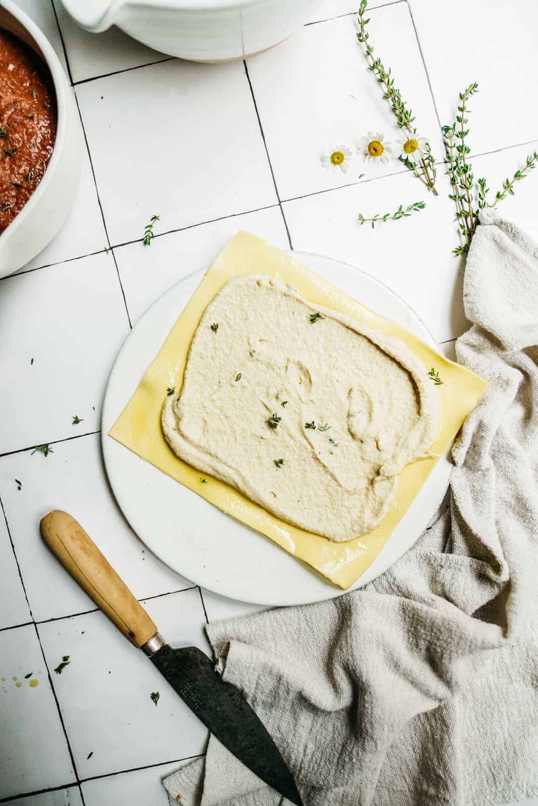 A plate with vegan lasagna roll ups topped with ricotta mixture and sprinkled with herbs sits on a white surface near a knife, a light cloth, scattered flowers, and thyme sprigs. A pot of red sauce is partially visible.