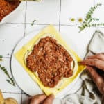 Hands assemble vegan lasagna roll ups, layering yellow pasta sheets with creamy white vegan cheese and rich tomato sauce on a white plate. Nearby are bowls of sauce, more cheese, fresh herbs, and a kitchen towel.