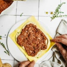 Hands assemble vegan lasagna roll ups, layering yellow pasta sheets with creamy white vegan cheese and rich tomato sauce on a white plate. Nearby are bowls of sauce, more cheese, fresh herbs, and a kitchen towel.