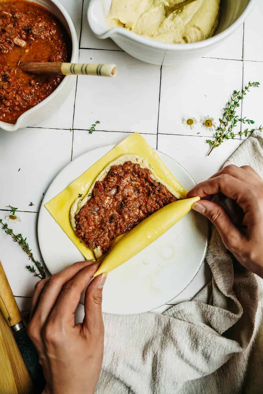 A person’s hands roll a sheet of pasta over a layer of sauce and vegan ricotta for vegan lasagna roll ups. Bowls of sauce, herbs, and a cloth are nearby on a white tiled surface.