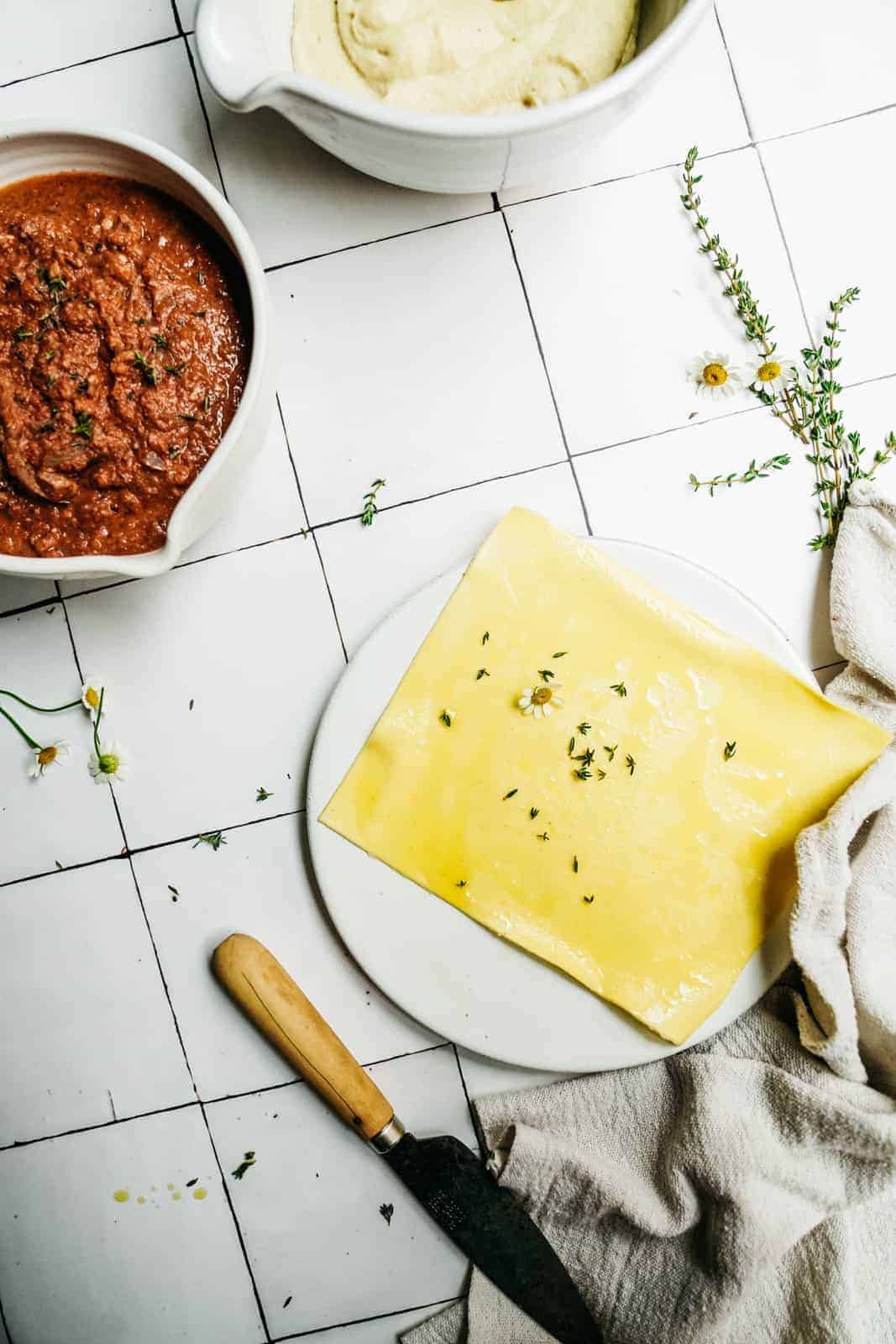 Overhead view of vegan lasagna roll ups on a white plate, sprinkled with herbs and a small flower, next to bowls of red tomato sauce and vegan ricotta, with a knife and a cloth on a tiled surface.