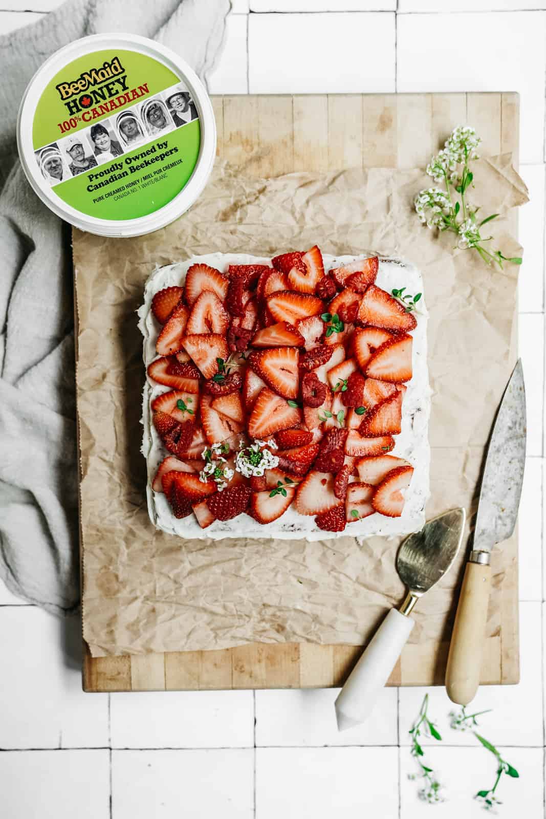 Bee Maid Honey sitting on cutting board with beautiful Honey Cake topped with strawberries.