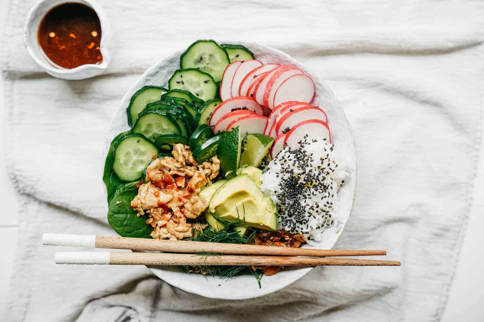 Vegan Tuna Poke Bowl sitting on white blanket with chilli sauce in background.