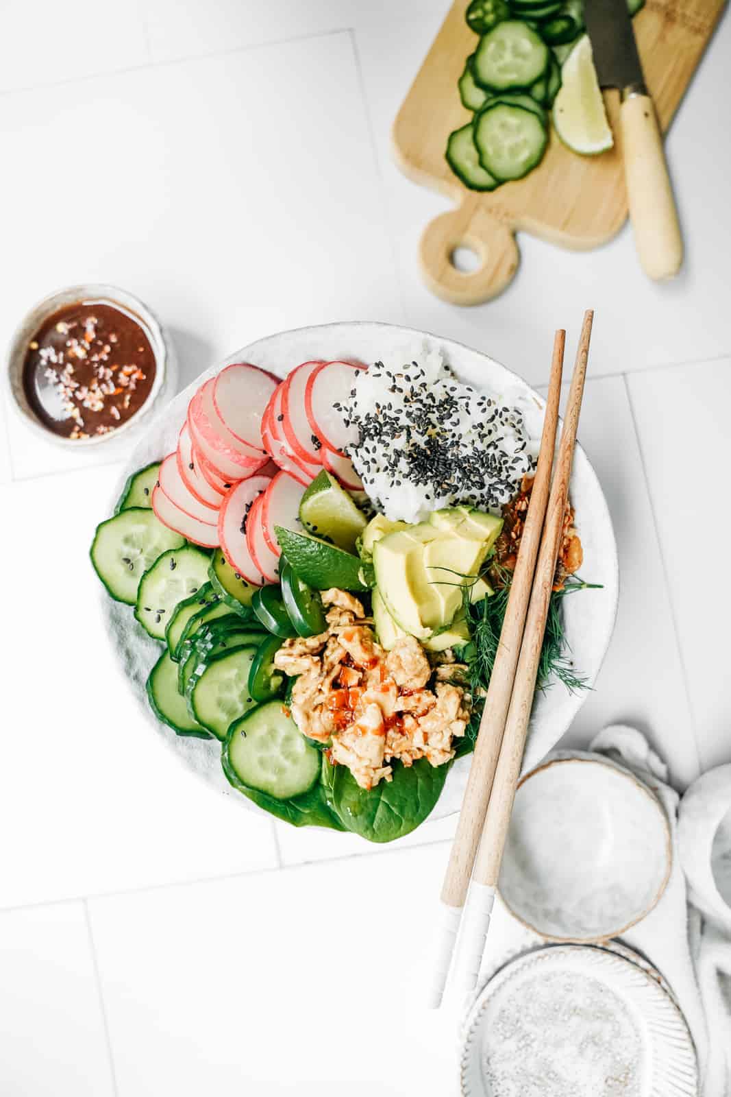 Vegan Poke Bowl sitting on table surrounded by ingredients like cucumber, spicy chilli sauce, and served with chopsticks.