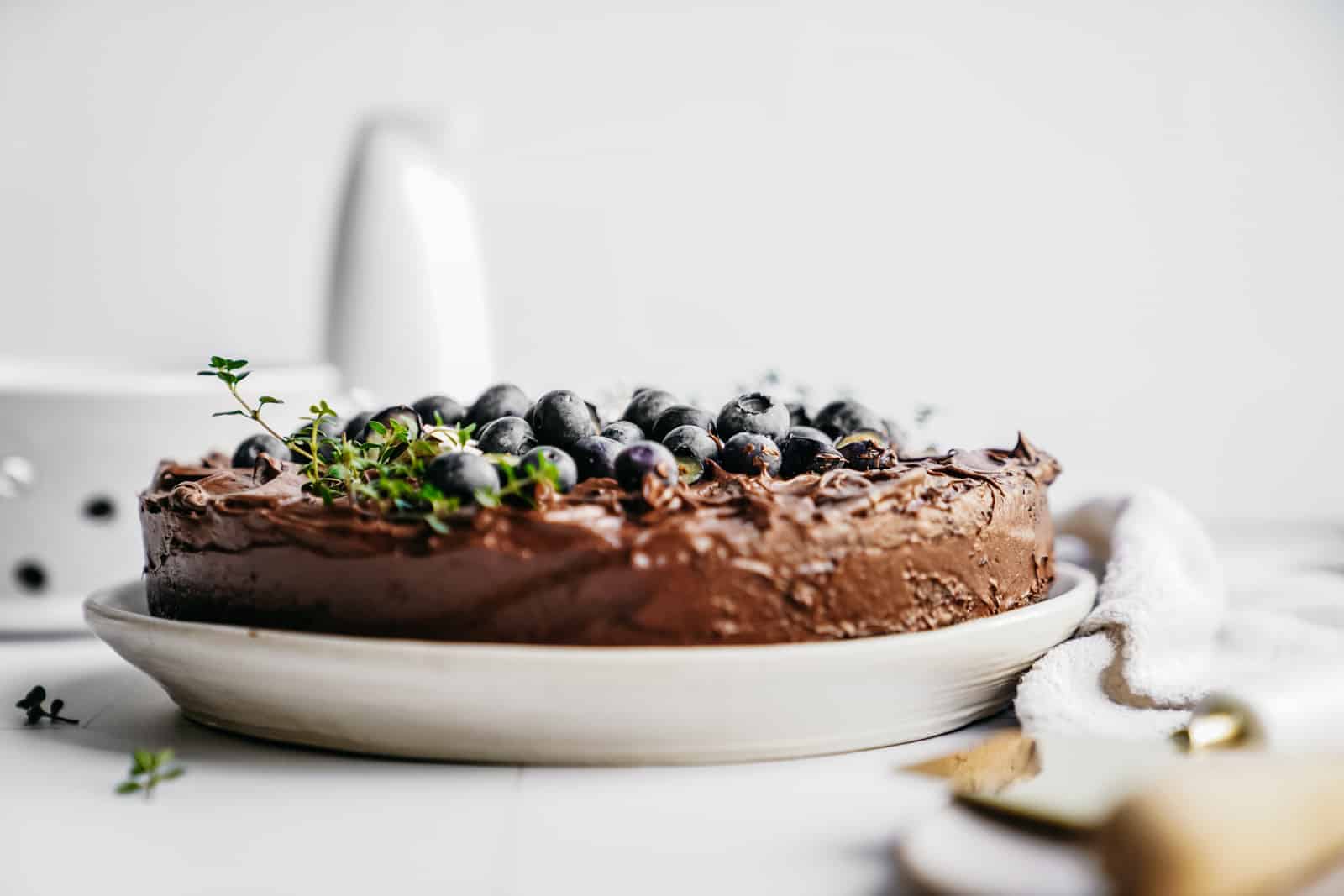 Side angle shot of a vegan chocolate cake that is topped with blueberries and fresh herbs.