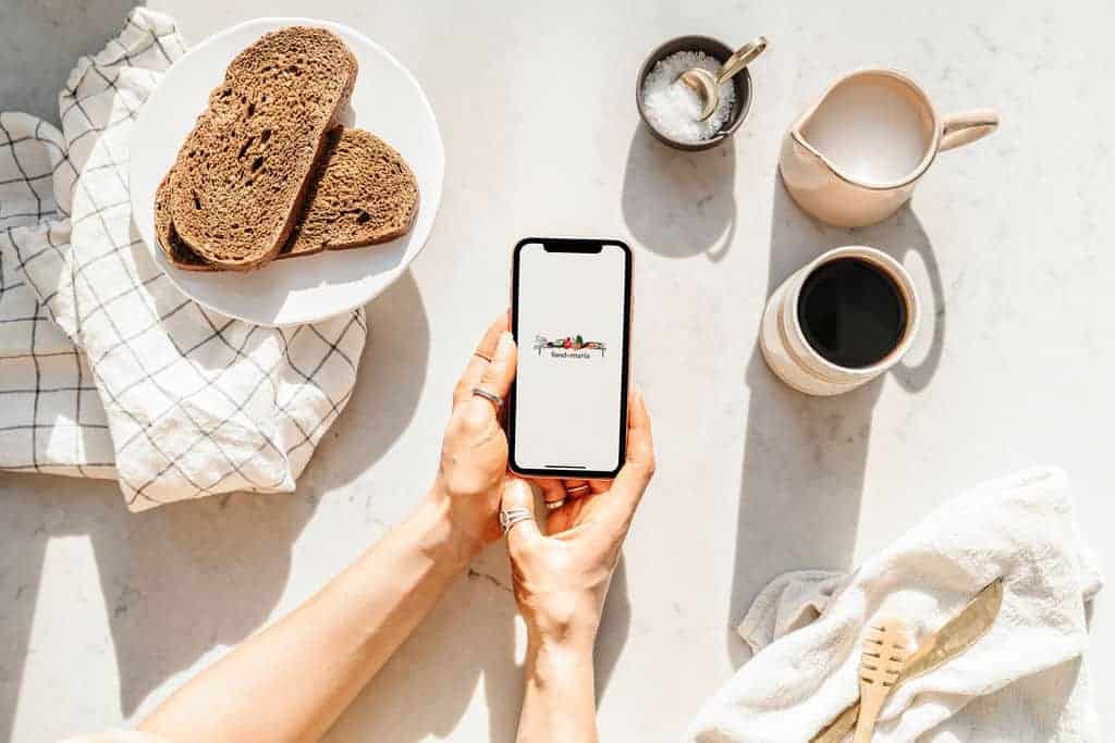 Hand holding a phone at a coffee table with breakfast displaying the FoodByMaria app, a great app for anyone, even food bloggers.