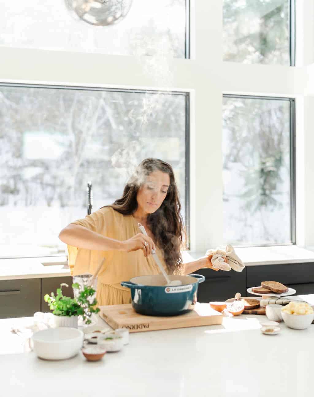 Maria stirring a pot of hot food at a white countertop with a beautiful winter backdrop