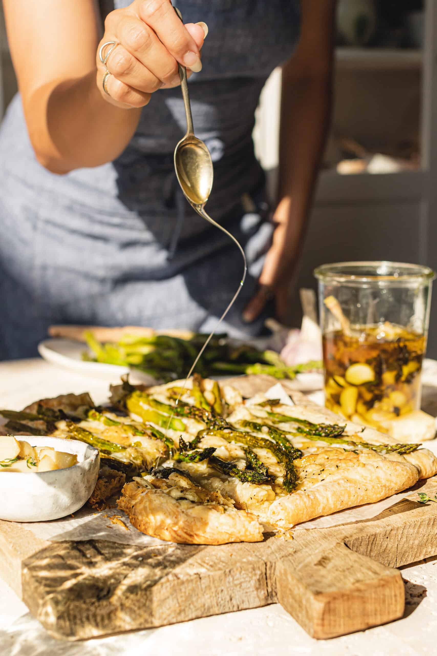 Drizzling honey on top of Asparagus Tart that is sitting atop a cutting board.