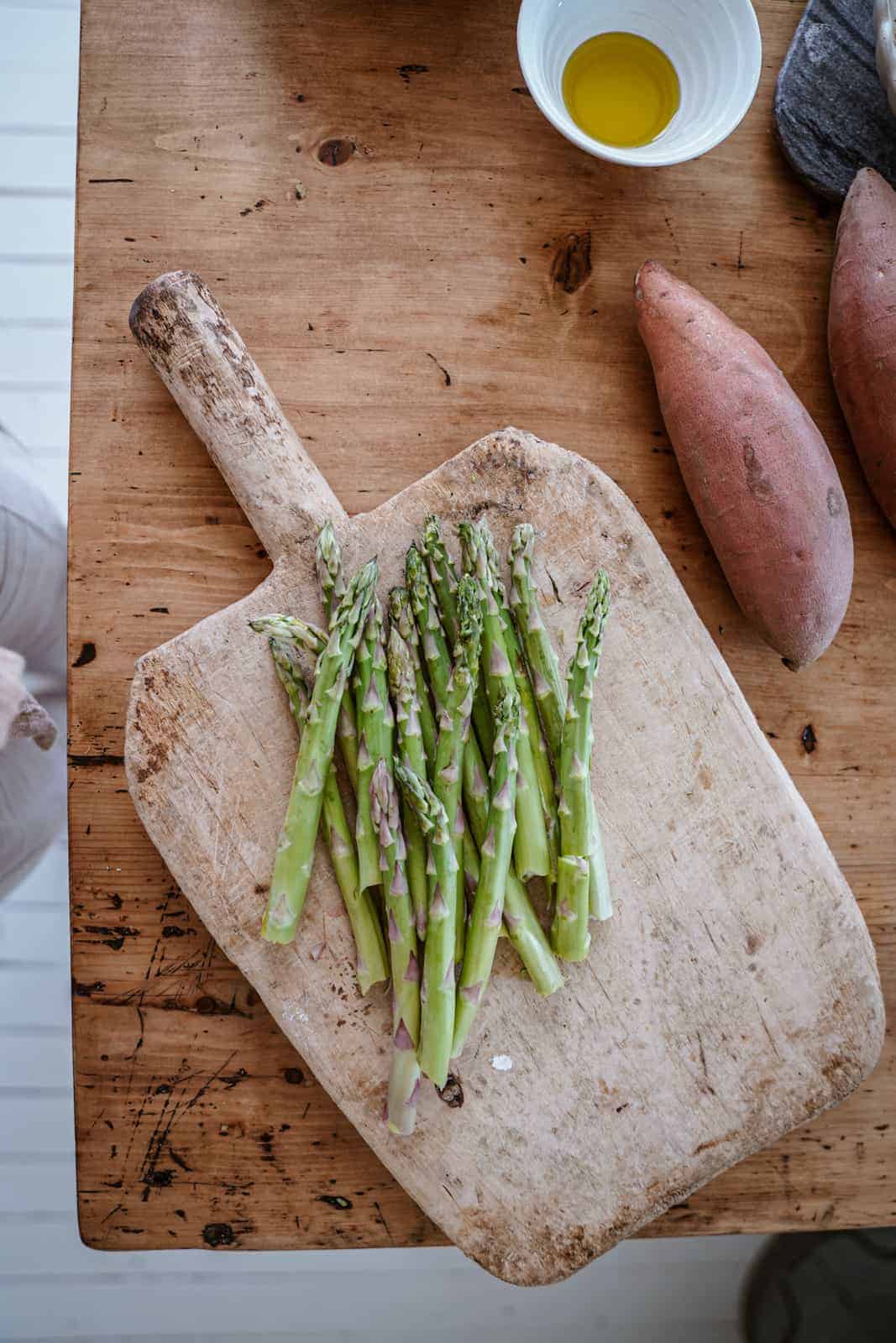 Asparagus on a cutting board surrounded by sweet potatoes.