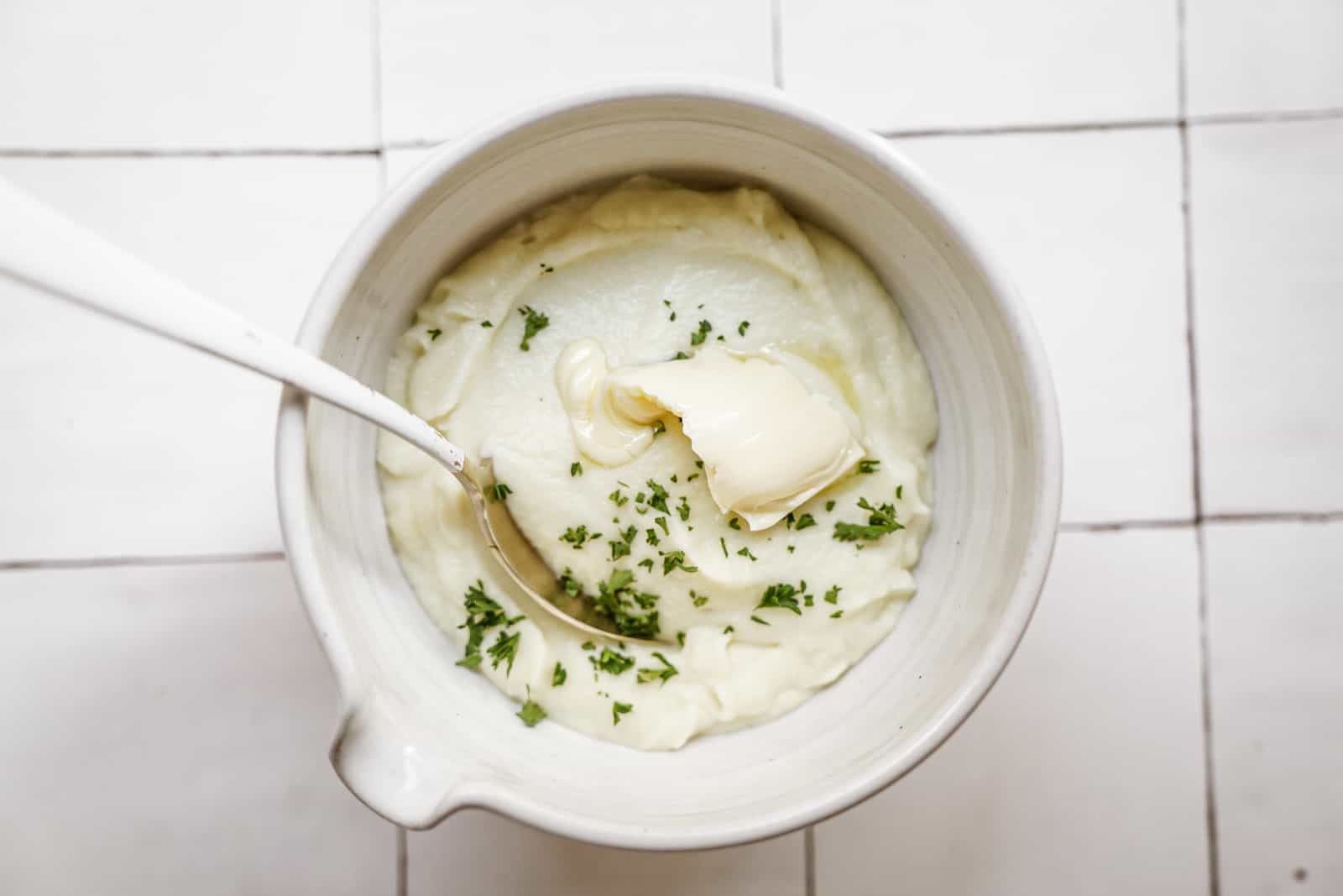 Vegan Mashed Cauliflower in a white bowl on white countertop with a spoon in it.