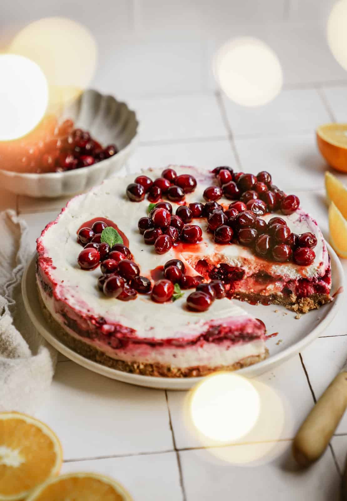 No-bake vegan cheesecake with cranberries on top, surrounded by oranges on countertop.