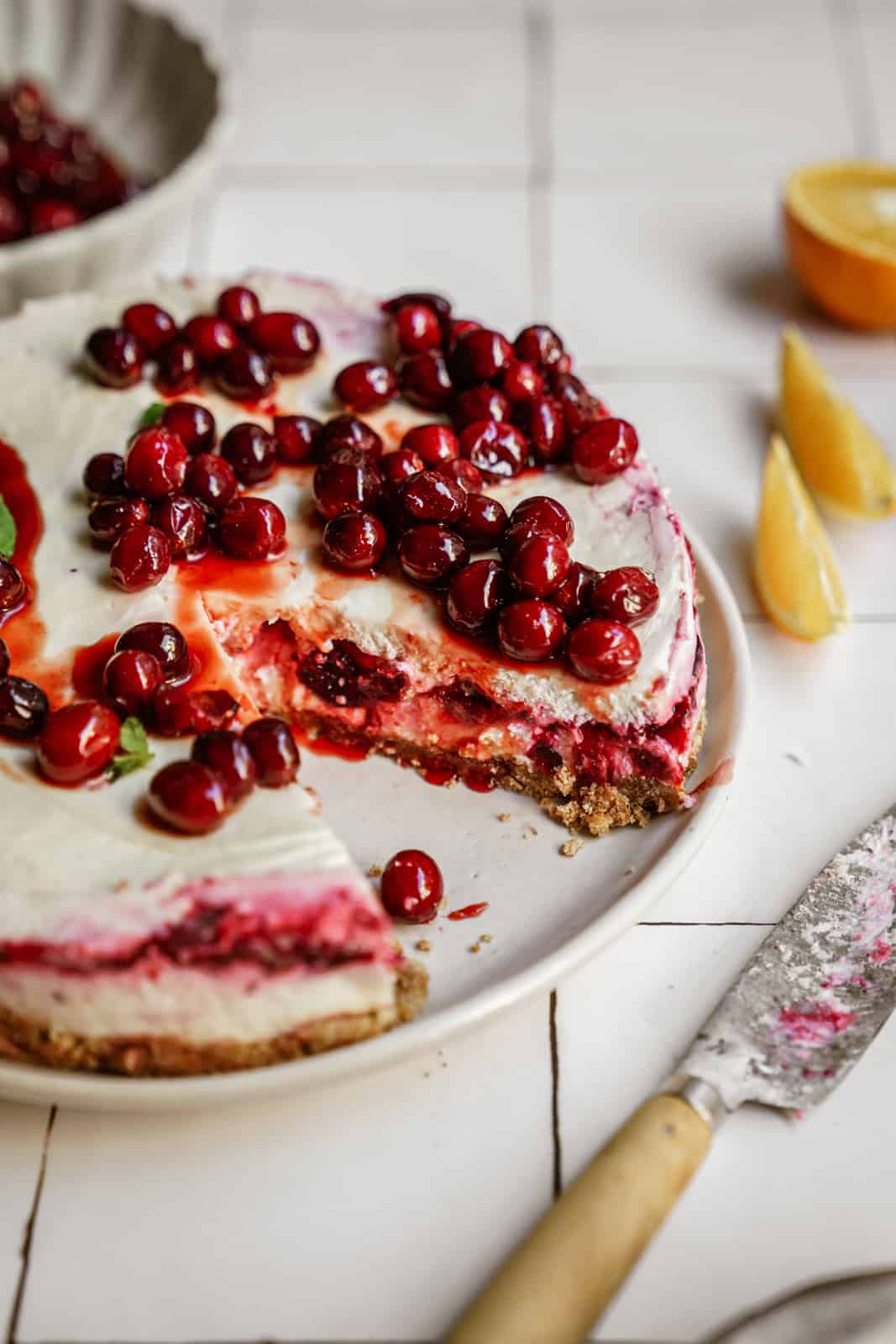 No-bake vegan cheesecake with cranberries and raspberries on top, surrounded by oranges on counter, with slice out of it.